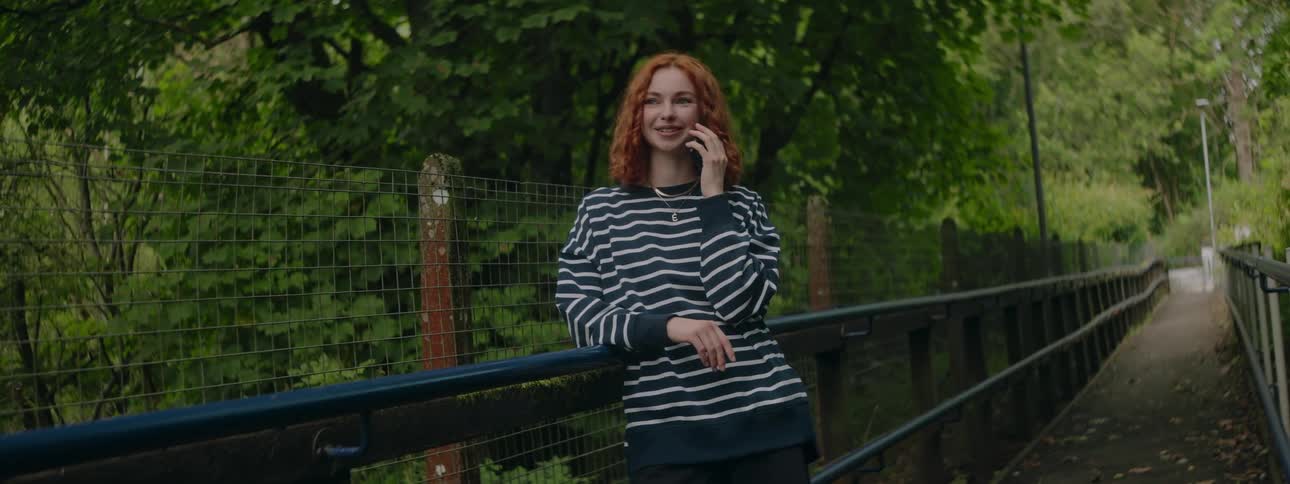 Redheaded woman talking on her cell phone while standing in a park surrounded by trees