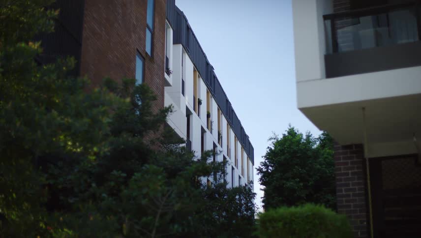 Two apartment buildings with brick and white facades separated by greenery against a pale blue sky