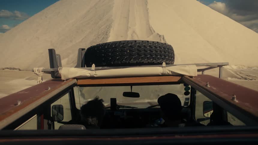 The roof of a car with the sunroof open with a tire on top and two people inside the car who are driving up a sand dune