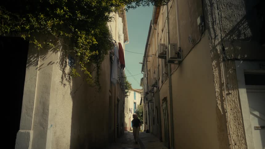 A narrow sunlit alleyway between tall weathered buildings with a lone figure walking towards the end of the street