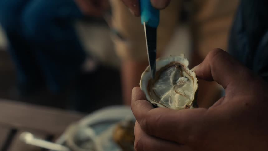 A person is holding an opened oyster in one hand and using an oyster knife in the other hand preparing it for consumption