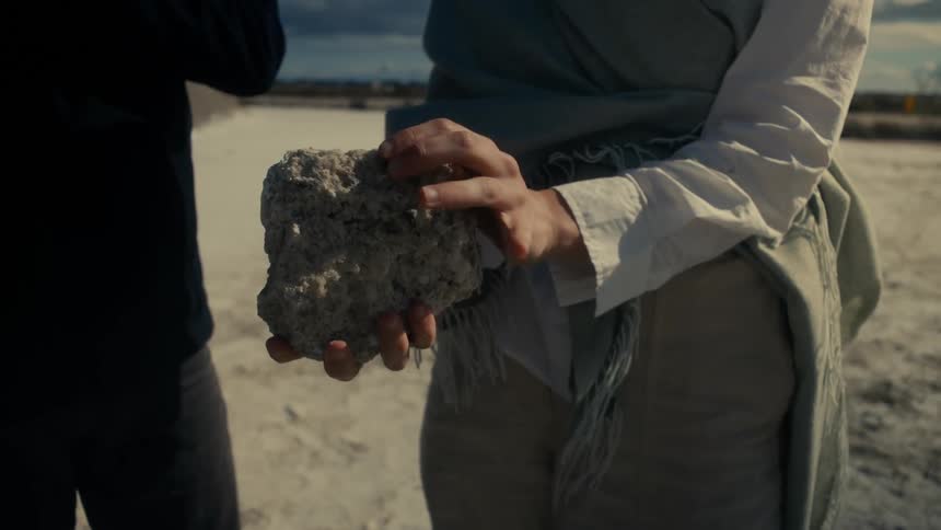 Woman stands in a sandy area with friends and turns over the rock she is holding
