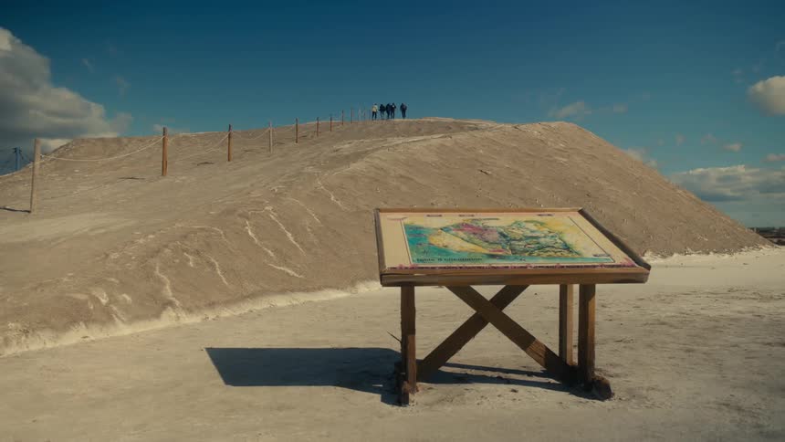 A large sandy hill with a wooden sign displaying a map a group of people stands at the top of the hill