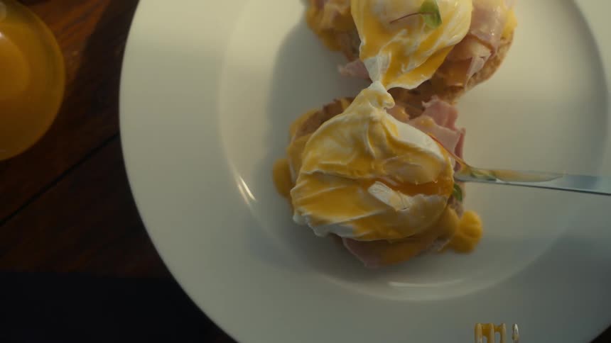 Plate of eggs benedict served on slices of ham and english muffins and a fork and knife are gently cutting through