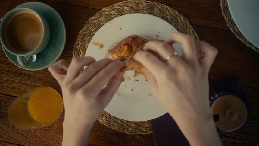A persons hands break apart a flaky croissant on a white plate with a cup of coffee and a glass of orange juice visible nearby