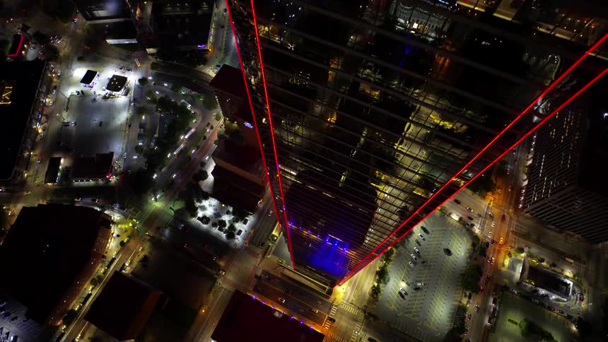An aerial view of a tall skyscraper illuminated with red lights surrounded by a grid of city streets and buildings