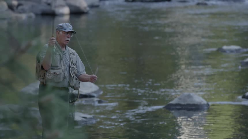 An older man is casting out his line as he fishes in a river