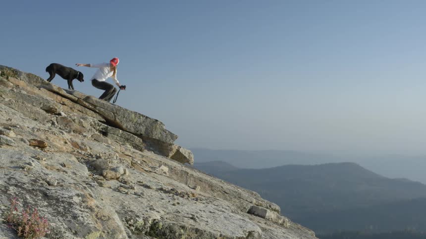 Woman standing on a slanted mountaintop as her dog slowly approaches to join her