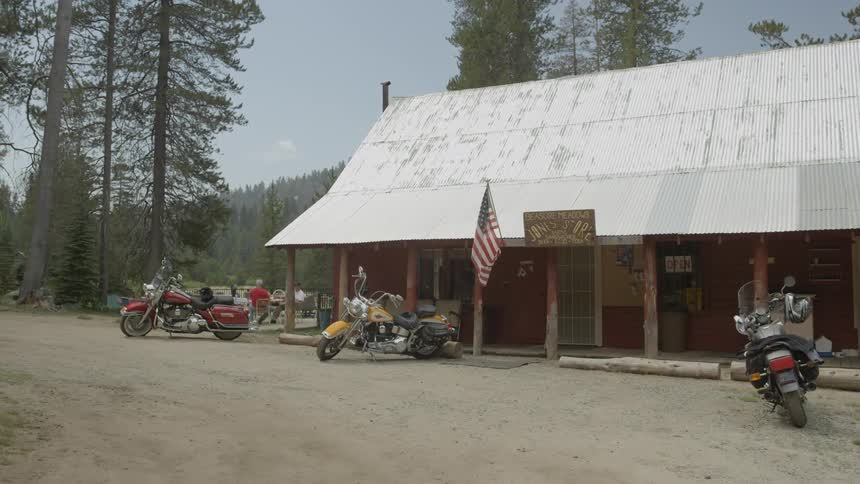 Motorcycles are parked outside an old roadside shop that's flying an american flag