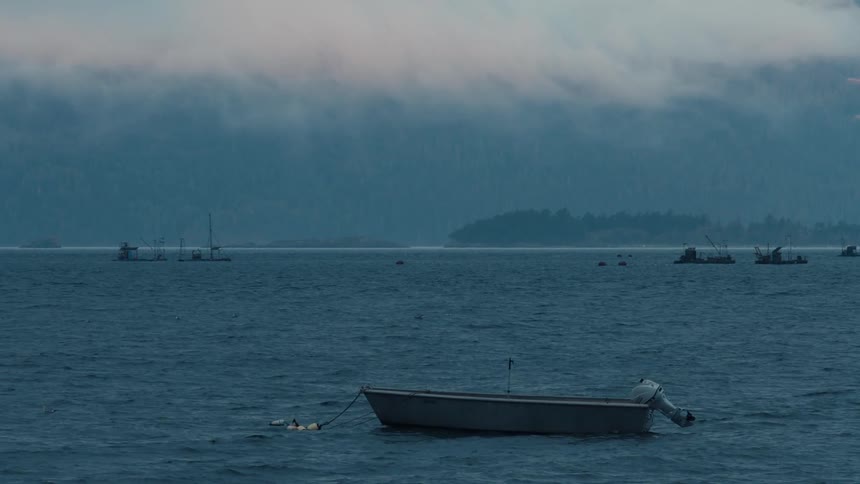 A small gray boat sits peacefully on a dark blue slightly choppy sea with a hazy overcast sky and other boats in the distance