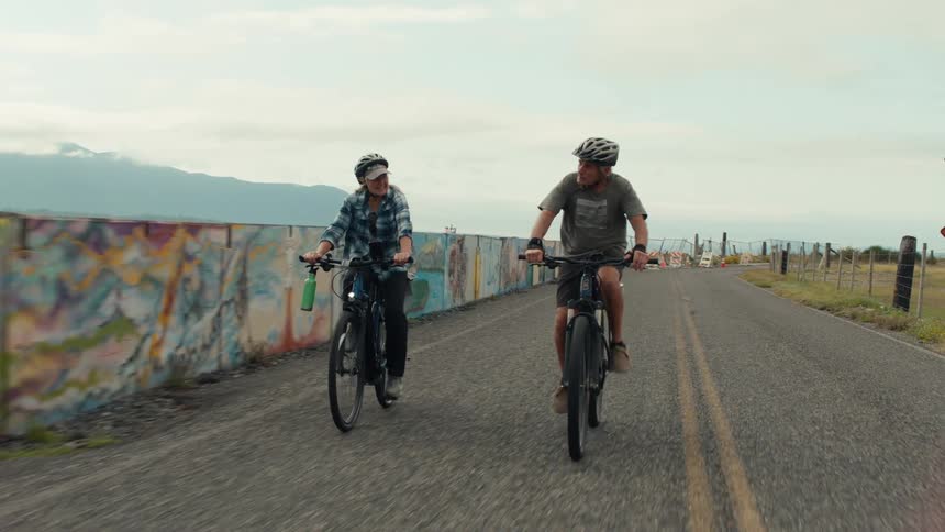 Two people wearing helmets ride bicycles on a paved road flanked by a colorful graffiticovered wall