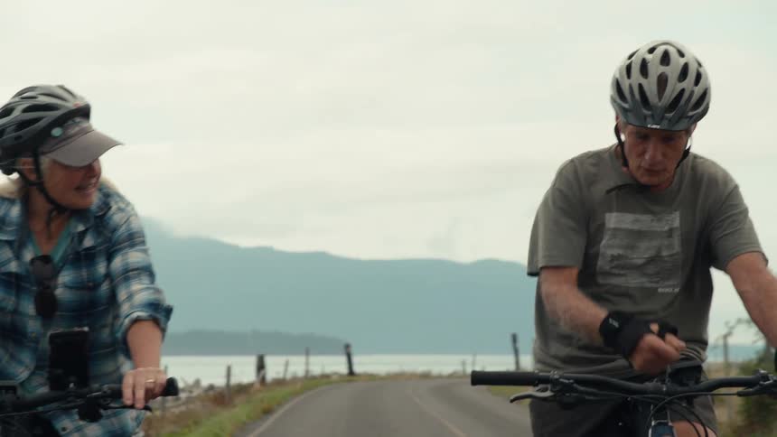 Two senior citizens ride bicycles on a road with a scenic background of mountains and water