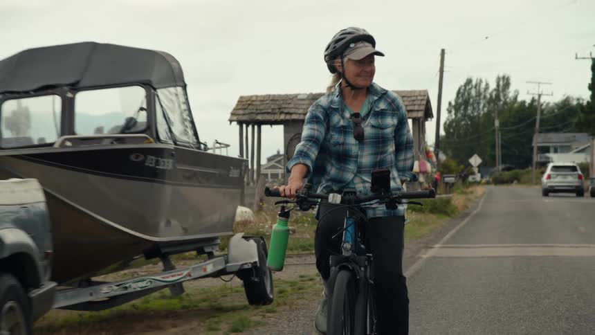 An elderly woman and a man ride bicycles past a parked truck towing a boat with a coastal town visible in the background
