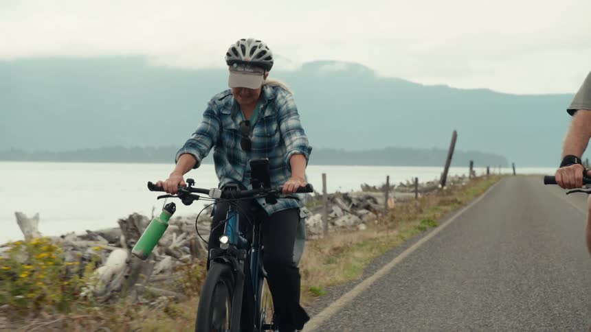 An elderly woman riding a bicycle with a green bottle hanging from her handlebars and she is on an asphalt street next to a large body of water