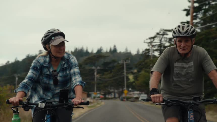 An elderly man and woman are riding bicycles happily wearing protective helmet on an asphalt street