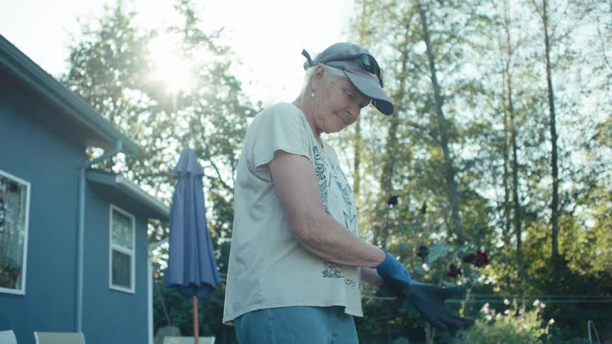 An elderly woman puts on gloves and starts gardening in her yard