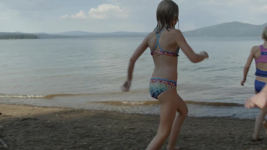 A group of young girls in bathing suits run into the water at a lake