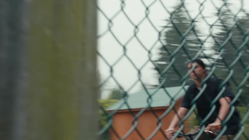 A man wearing a black shirt brown pants and a black backpack rides a bicycle through a chainlink fence disappearing into a green expanse