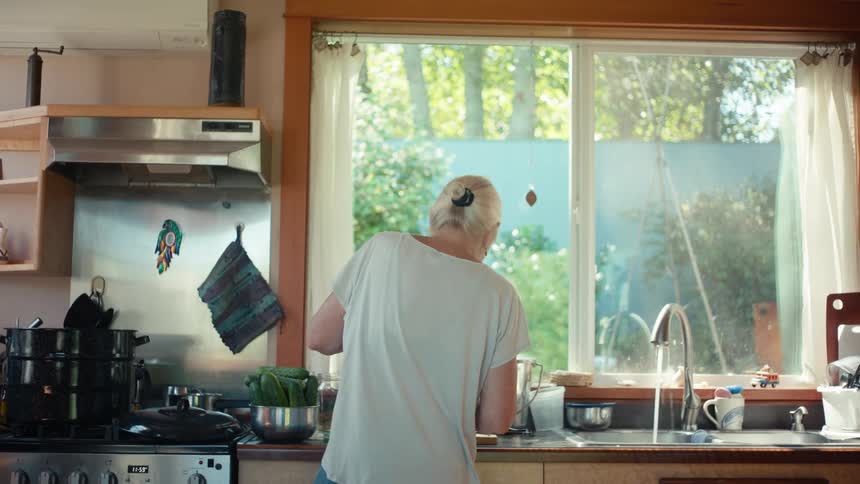 An older woman with white hair in a bun standing in a cozy kitchen facing a large window with sunlight streaming in overlooking a garden