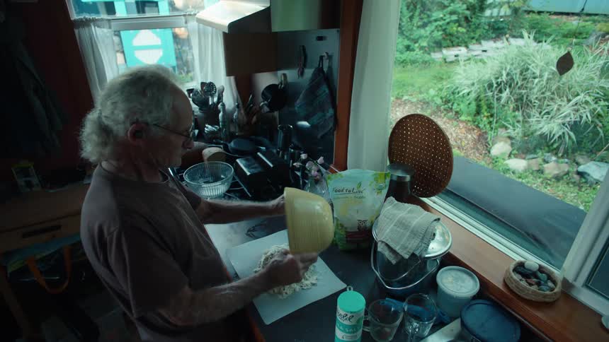 An elderly man kneads dough on a kitchen counter with a scenic garden visible through the window