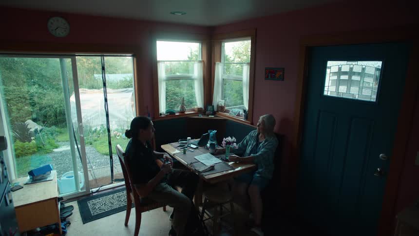 Two people sitting at a wooden table in a cozy room with large windows having a conversation while sunlight illuminates a garden visible through a sliding glass door