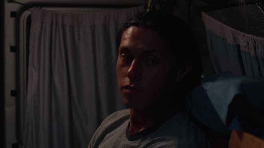 A person sits resting inside a vehicle with curtains wearing a gray shirt