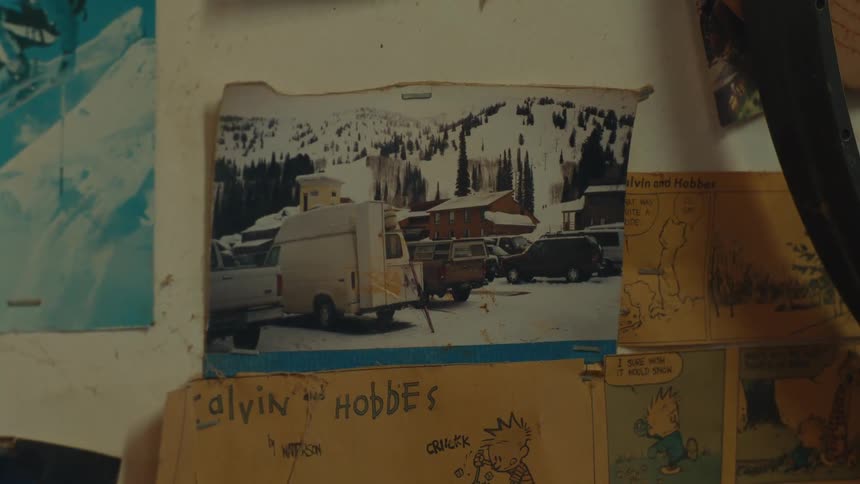 An old photo of a snowy mountain landscape with a cabin and cars sits on a wall surrounded by other photos and drawings