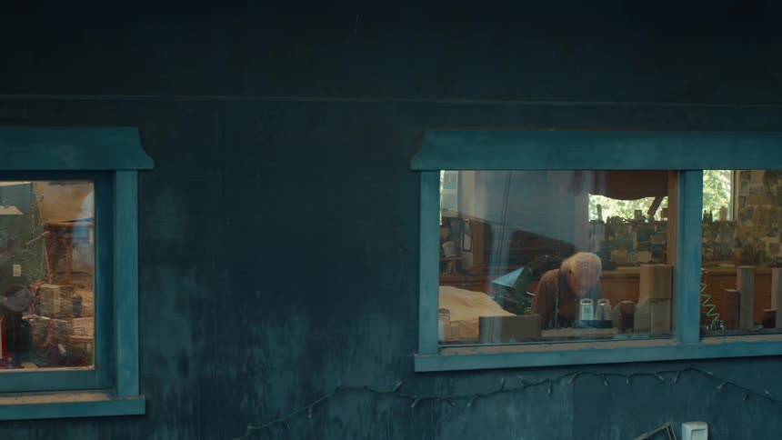 A man works at a workbench inside a dimly lit workshop seen from a window