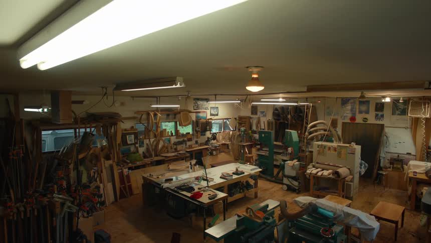 The fluorescent lights flicker on illuminating a cluttered workshop filled with woodworking tools and unfinished projects