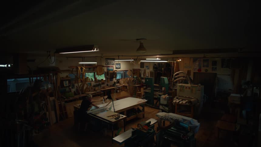 A dimly lit workspace with a large table covered in tools and fabrics and a person sitting at the table working on a project