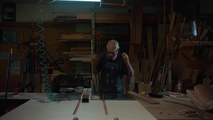 An older man working in a dimly lit woodworking shop surrounded by tools and wooden planks intently focusing on a task at a central workbench