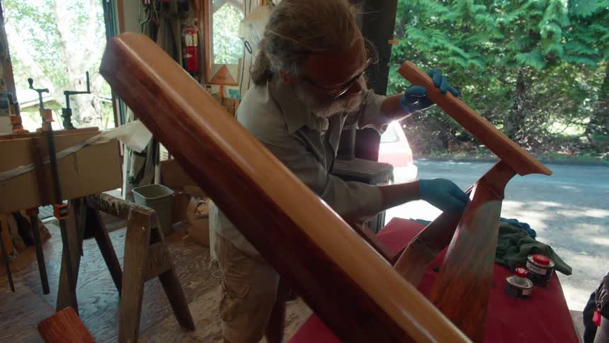 A grayhaired man wearing glasses and blue gloves is sanding the surface of a wooden piece of furniture