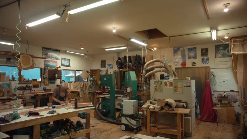An elderly man in a green apron is fiddling with wooden object in a factory full of scattered tools