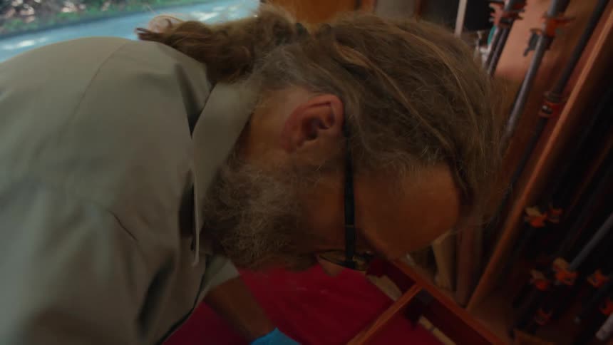 An older man is closely focused on applying a finish to a wooden chair in a workshop