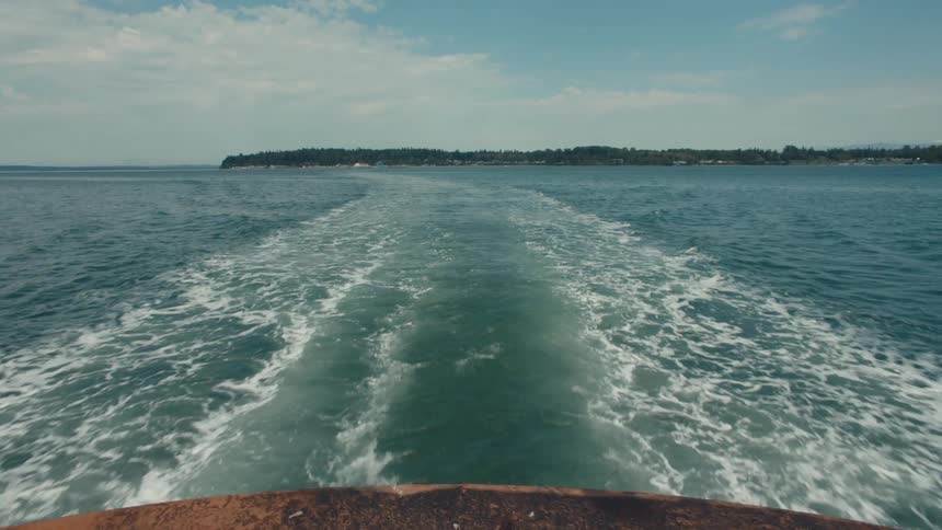 A boat leaves a trail of rippling waves across a calm blue sea heading away from a distant shoreline