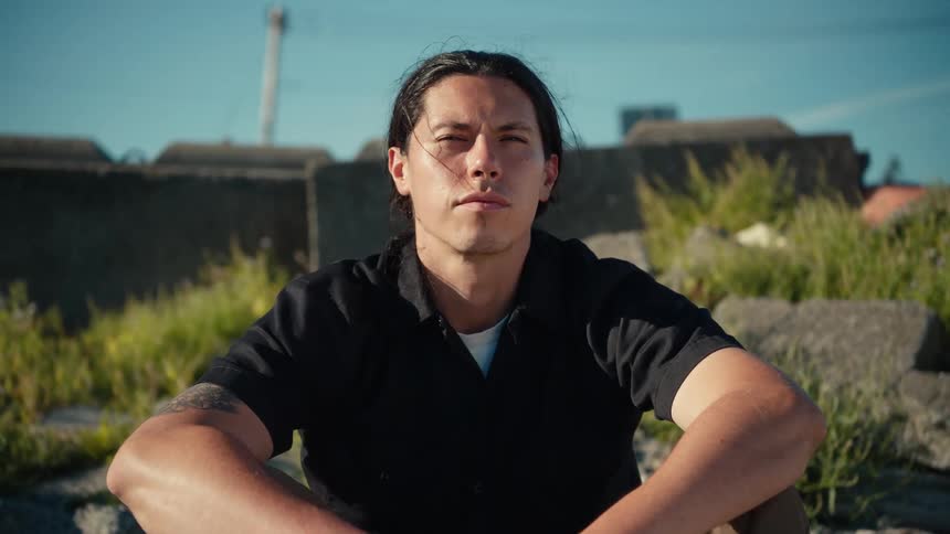 A man with long hair sitting outdoors on a rocky surface under clear skies with overgrown grass and a concrete wall in the background conveying a calm and contemplative mood