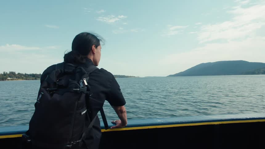 A person with a backpack gazes at the calm waters and distant hills from a boat