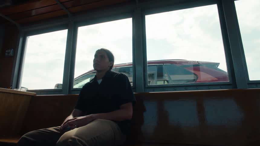 A man is sitting indoors on a bench that is next to windows where there is a car outside and the man looks to his left side