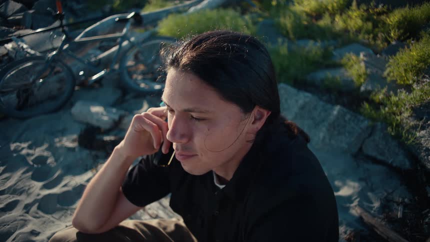 A rusted bicycle lies abandoned on a sandy shore littered with debris and large rocks while a man with long hair wearing a black shirt sits nearby engrossed in a phone conversation