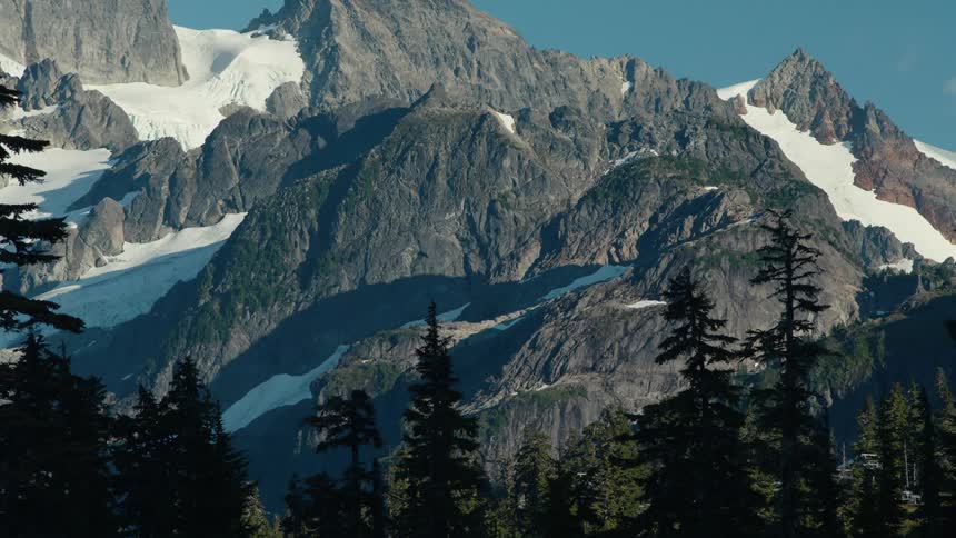 A huge stone mountain with some snow on its surface and a pine forest in front