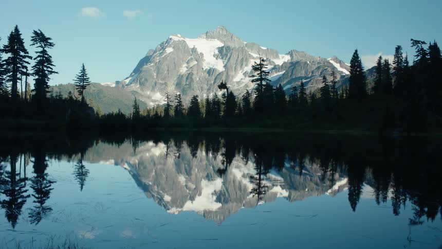 A snow capped mountain reflects perfectly in a still lake surrounded by a forest of tall trees