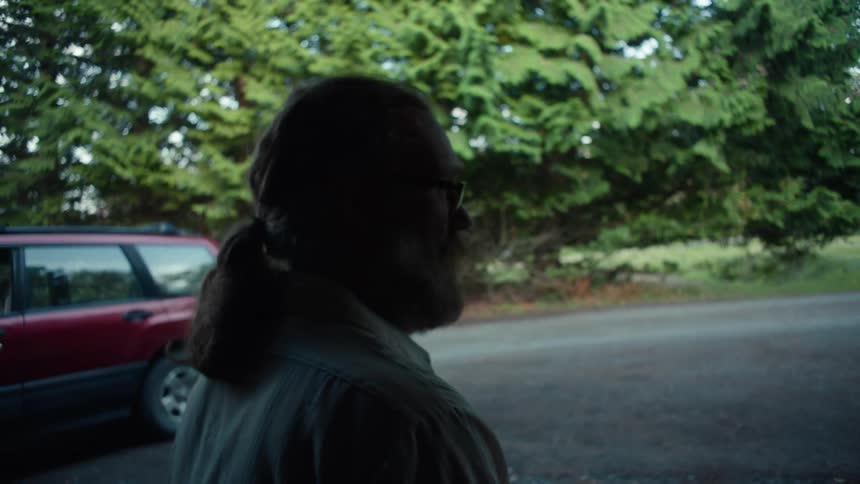 A bearded man with long hair and a shirt talks to person near a red car and green forest