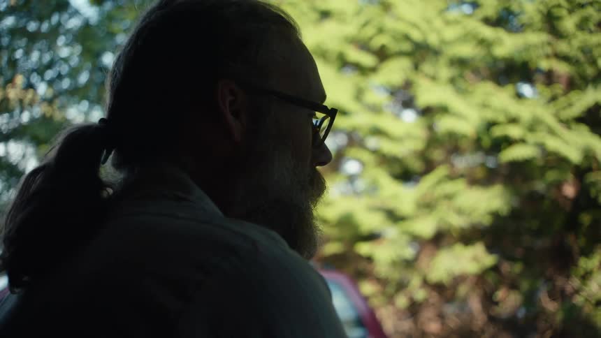 The face of a man with a gray beard and wearing glasses who is talking to someone who is in front of him and there are some green leaf trees behind