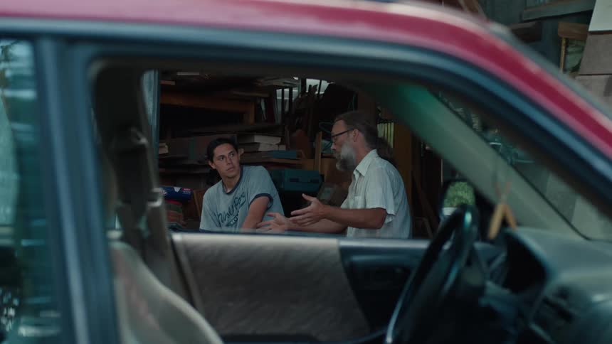 Two people in a workshop are viewed through a vehicles open window one person gestures while the other listens