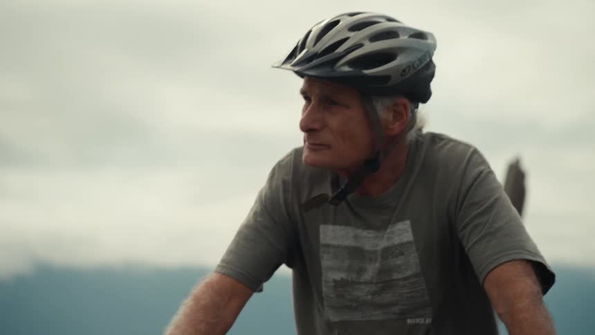 Two people are cycling along a road next to a body of water with mountains in the background