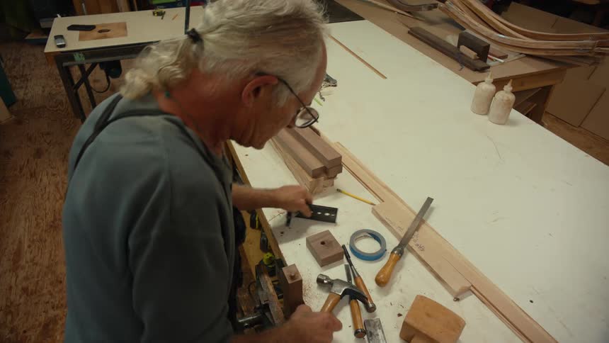 An elderly man and carpenter is using a hammer to nail a tool into a piece of wood