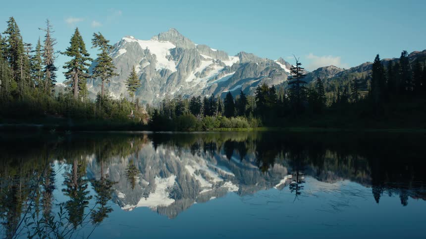 Calm lake reflecting its surroundings with conifers and a snowy mountain in the background