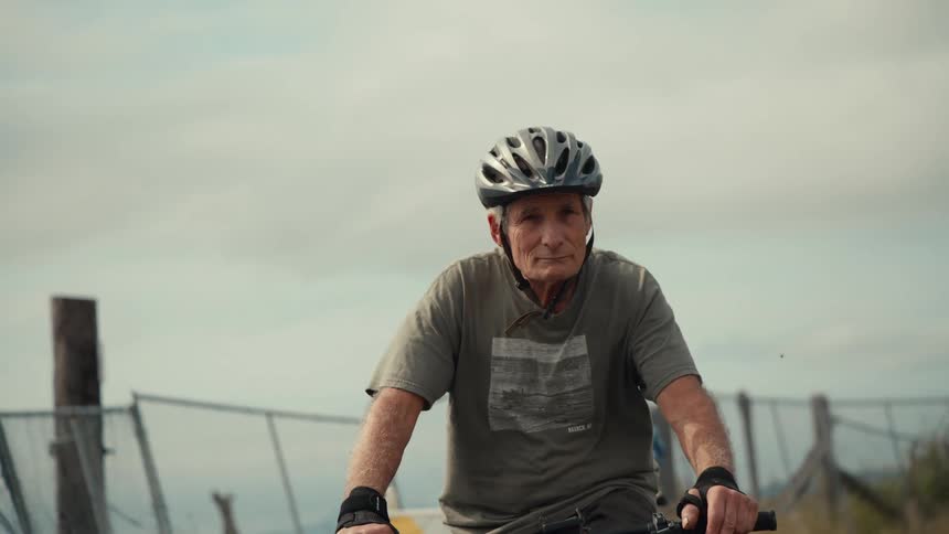 An older man and woman ride bicycles next to a fence near a river