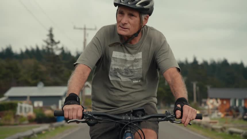 An elderly man and woman wearing helmets riding bicycles on a paved street in a neighborhood full of houses