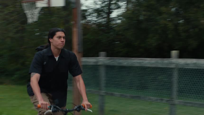 A man riding a bicycle on a basketball court surrounded by a parklike setting with trees and greenery in the background